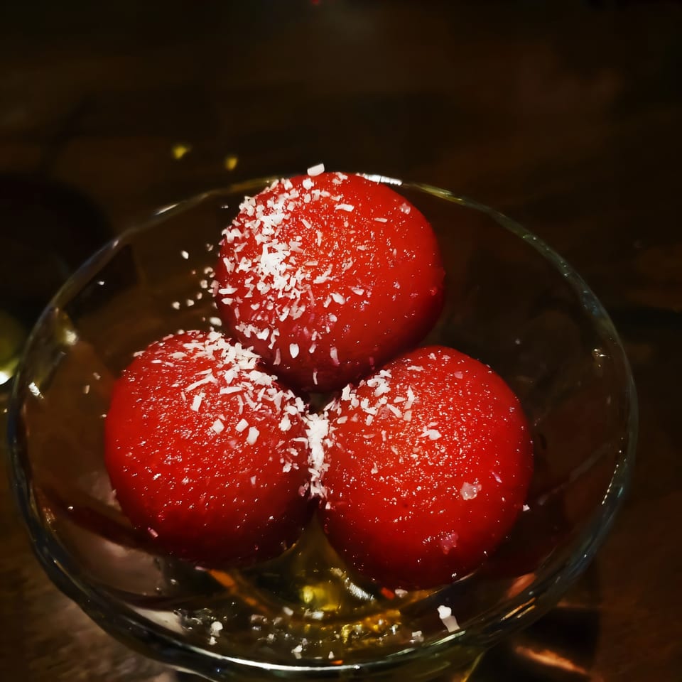 Three red gulab jamun, sprinkled with coconut flakes, in a clear glass bowl.