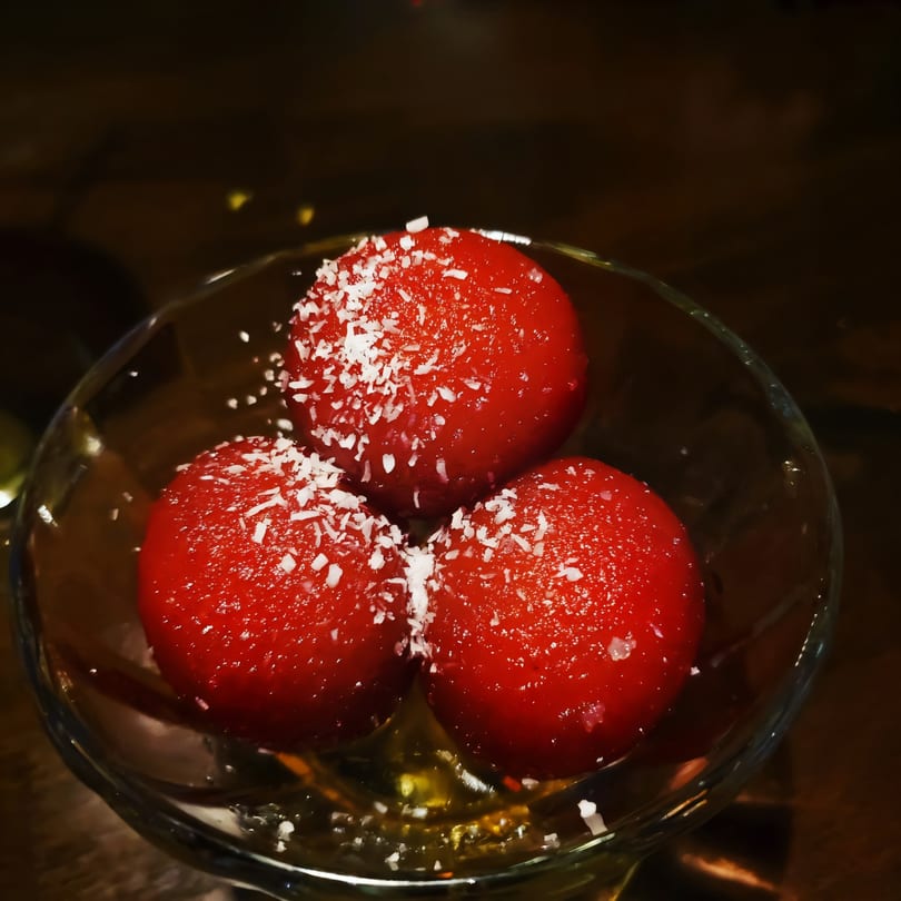 Three red gulab jamun, sprinkled with coconut flakes, in a clear glass bowl.