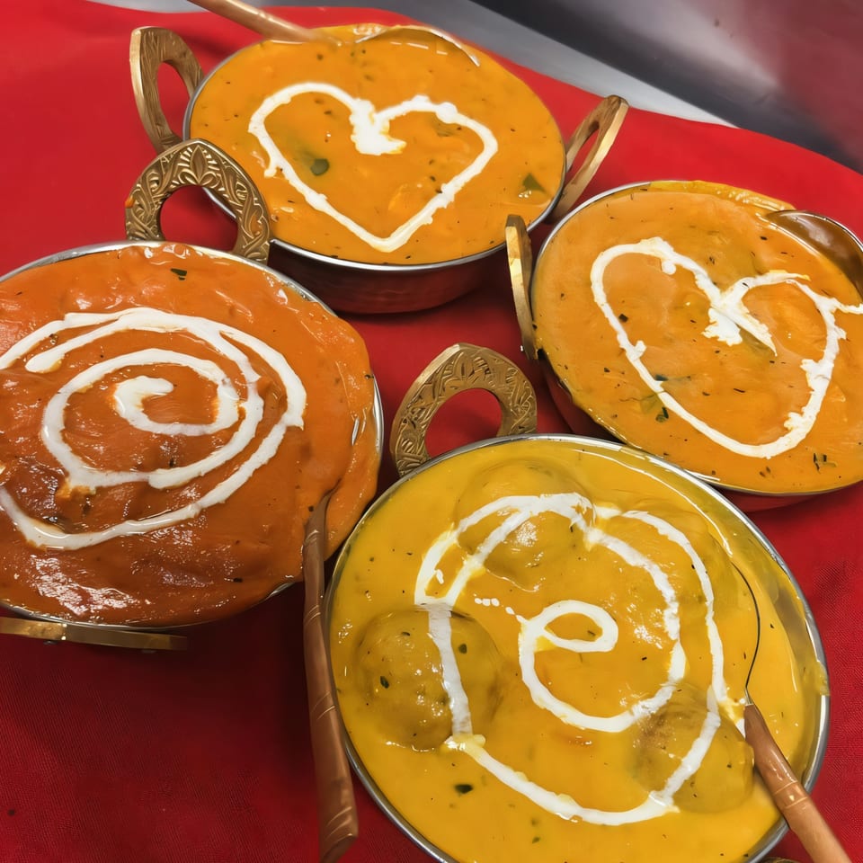 Four bowls of Indian curry, two orange and two yellow, with cream swirls on a red tablecloth.